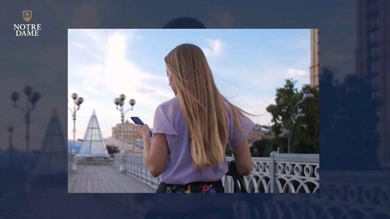 TBlond woman in purple top and floral skirt walks on a bridge, looking at her blue smartphone. Modern glass pyramids in background.